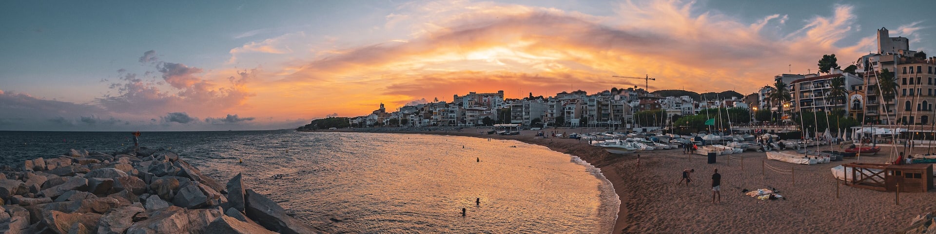 Una playa con costa rocosa, barcos y edificios al fondo durante el atardecer.