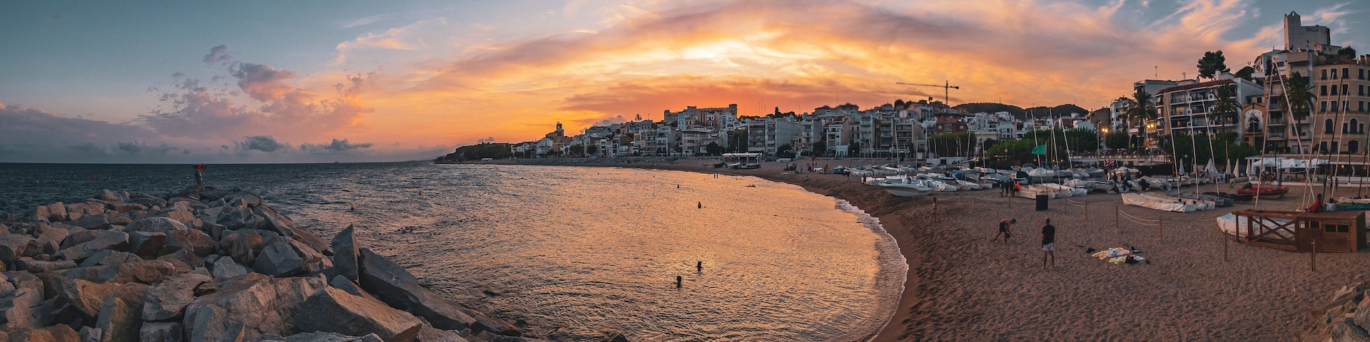 Una playa con costa rocosa, barcos y edificios al fondo durante el atardecer.