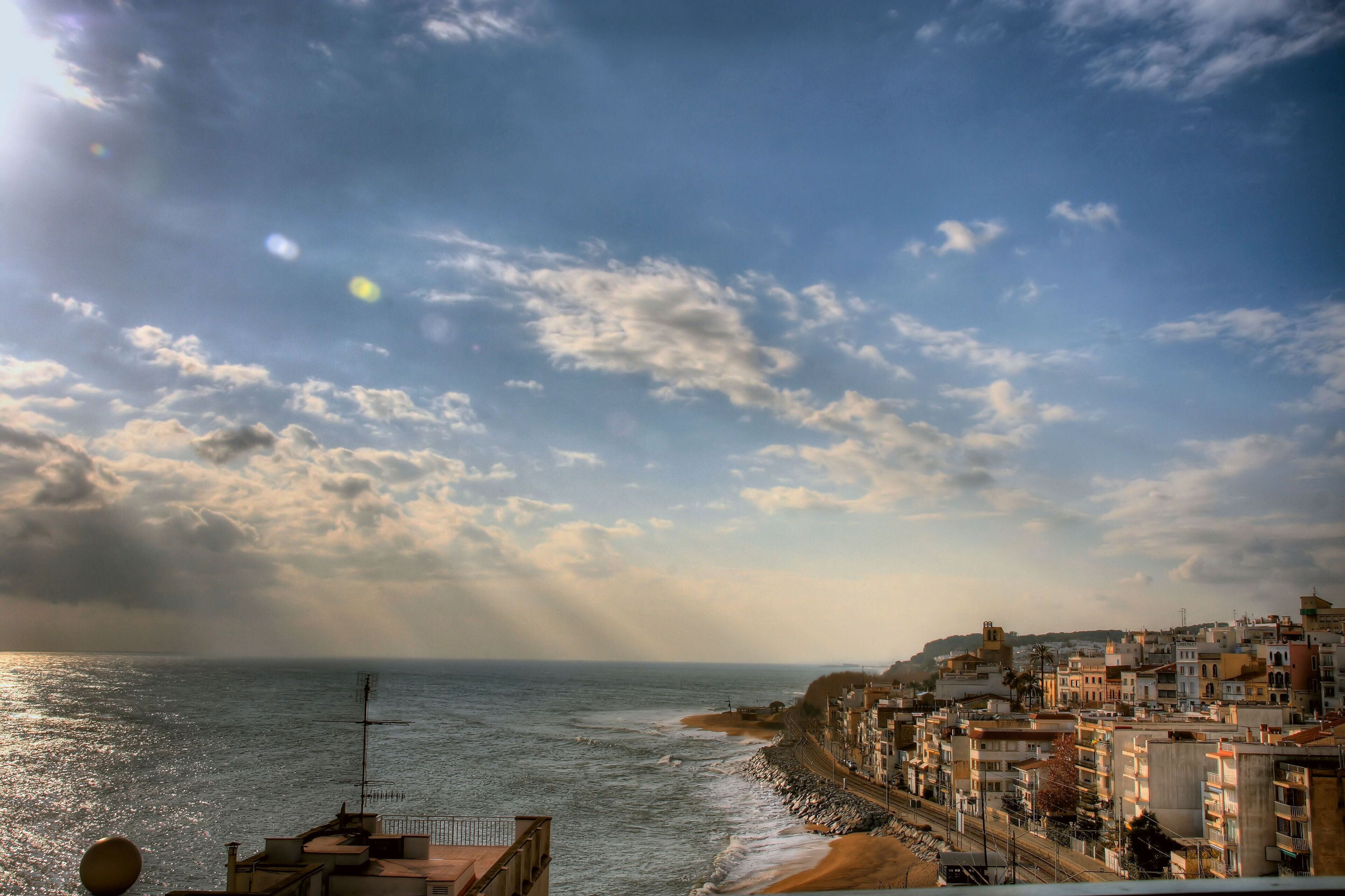 A coastal cityscape with buildings along the shore, a beach, and a cloudy sky.