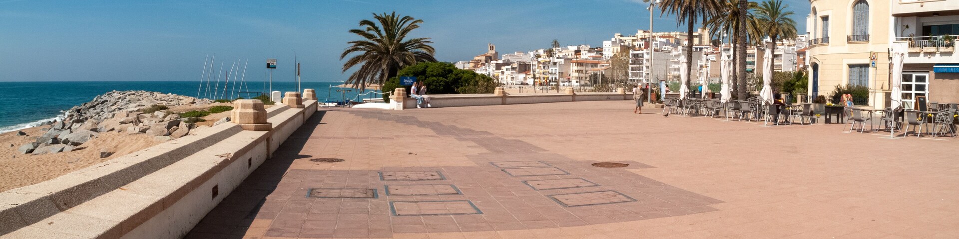 A coastal promenade with a sandy beach, palm trees, and a row of buildings.
