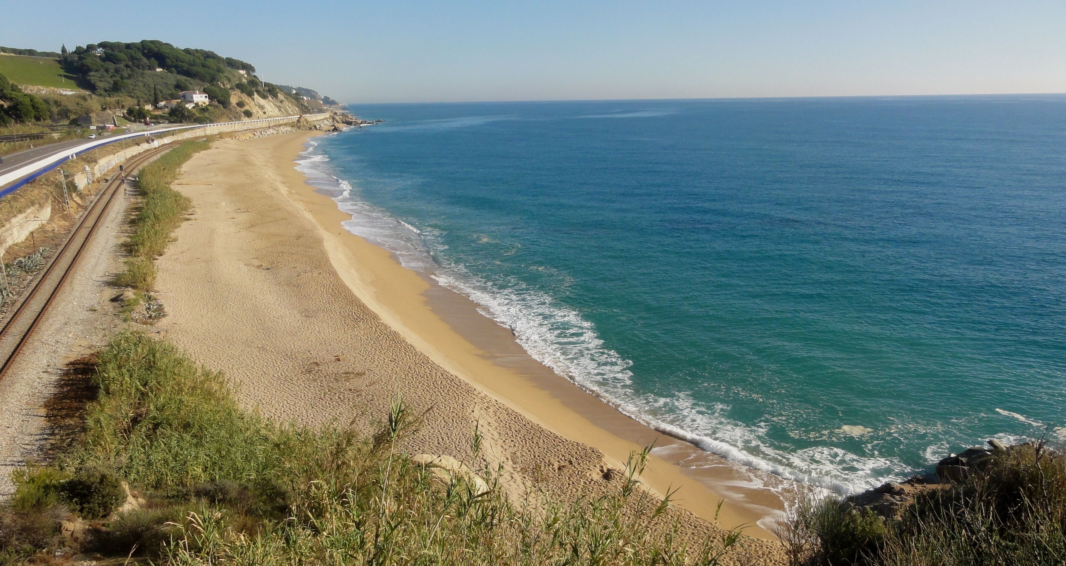 A coastal landscape with a sandy beach, a railway track parallel to the shore, and a clear blue sea.