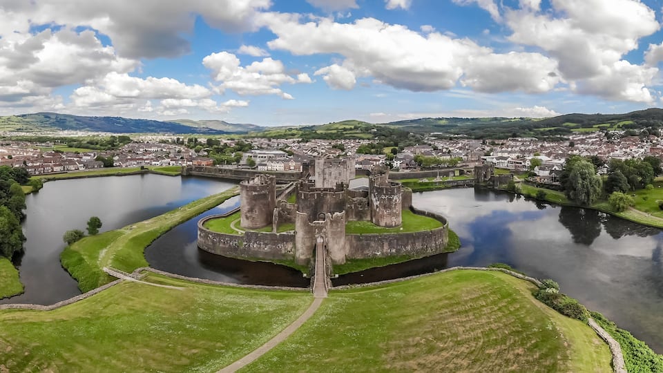 A historic castle surrounded by a moat, a town in the background, and rolling hills.