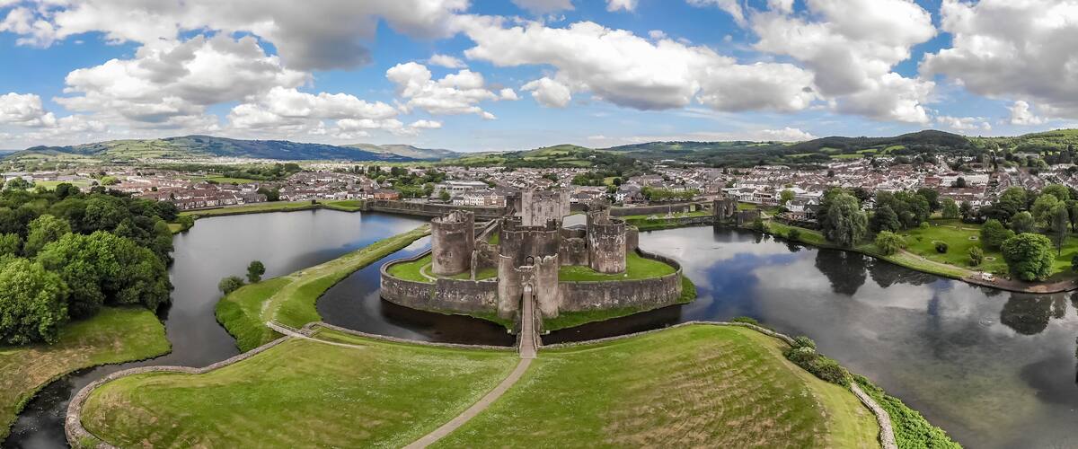 A historic castle surrounded by a moat, a town in the background, and rolling hills.
