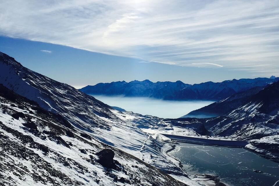 Paisagem de montanha coberta de neve, com um lago e montanhas ao longe, sob um céu claro.