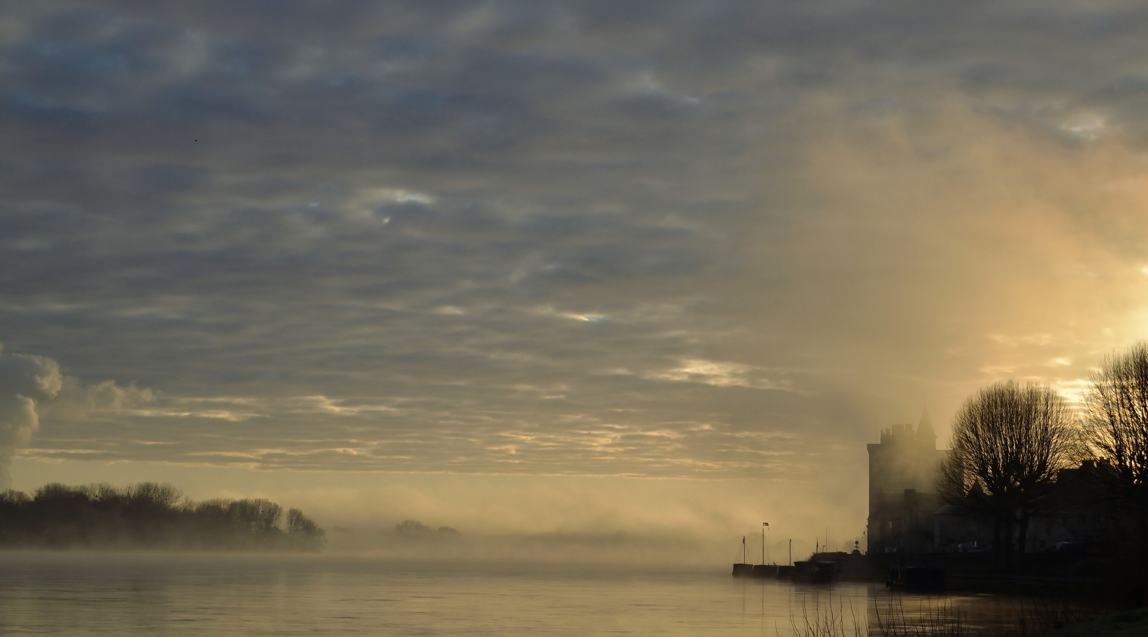 A river with a misty landscape, a distant city skyline, and a few boats.