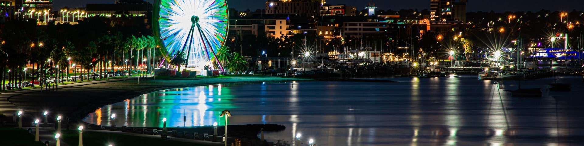 Un paisaje urbano frente al agua con una noria iluminada, un puerto deportivo con barcos y un puente sobre el agua.
