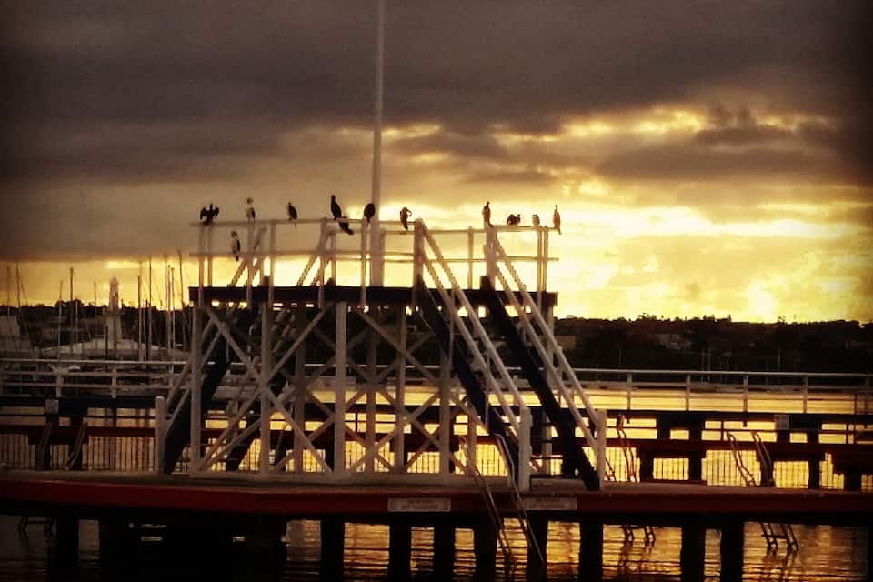 A pier with a metal staircase and railings, boats docked in the background, and a cloudy sky.