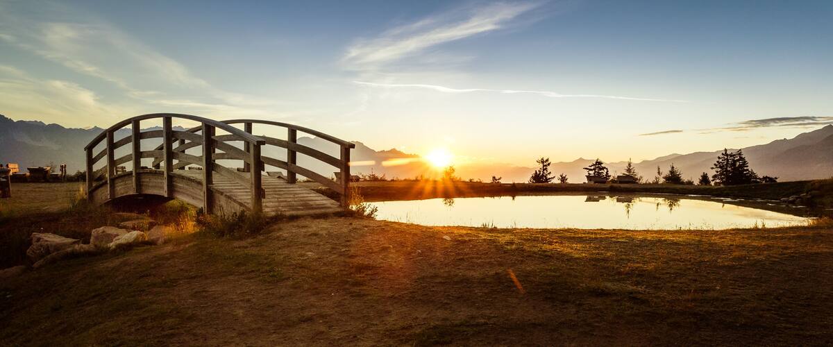 A wooden bridge over a calm lake with mountains in the background and the sun setting.