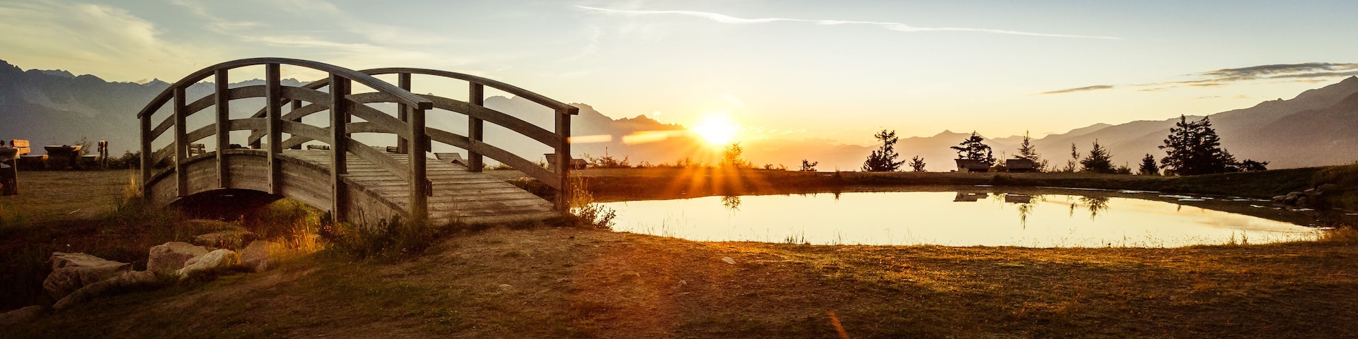 A wooden bridge over a calm lake with mountains in the background and the sun setting.