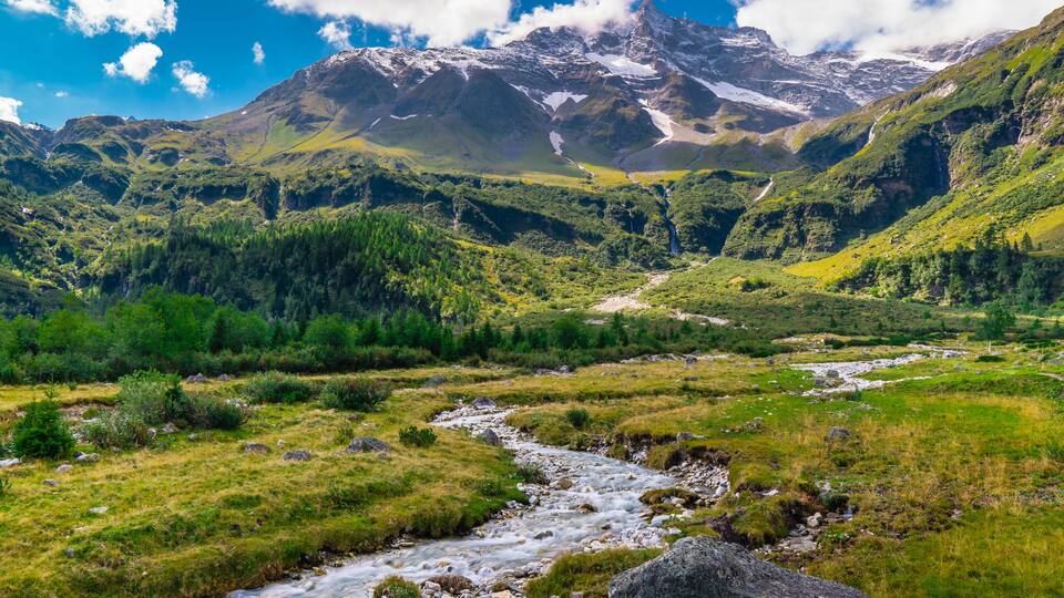 A mountain landscape with a river flowing through a valley, surrounded by lush greenery and snow-capped peaks.