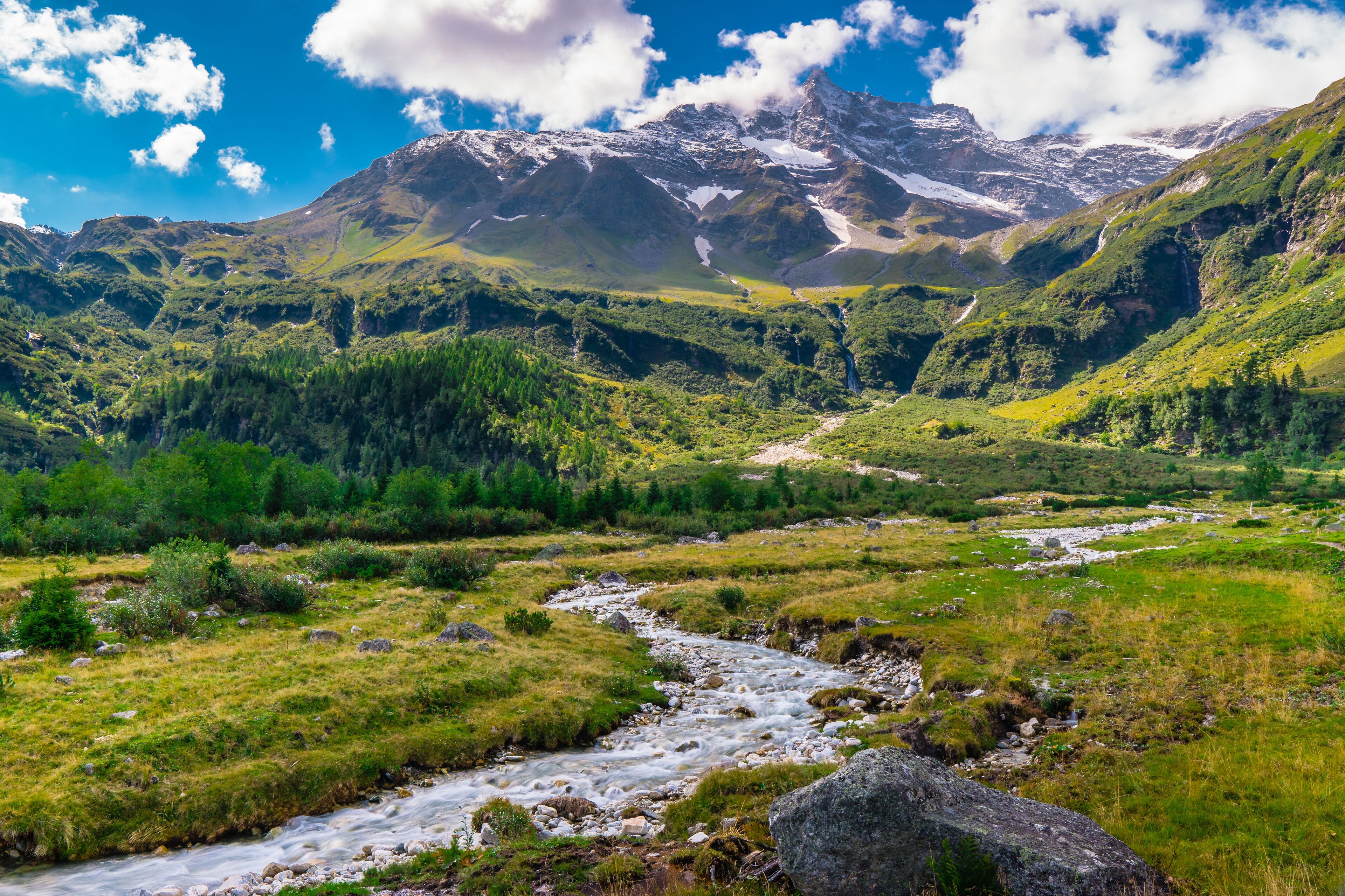 A mountain landscape with a river flowing through a valley, surrounded by lush greenery and snow-capped peaks.