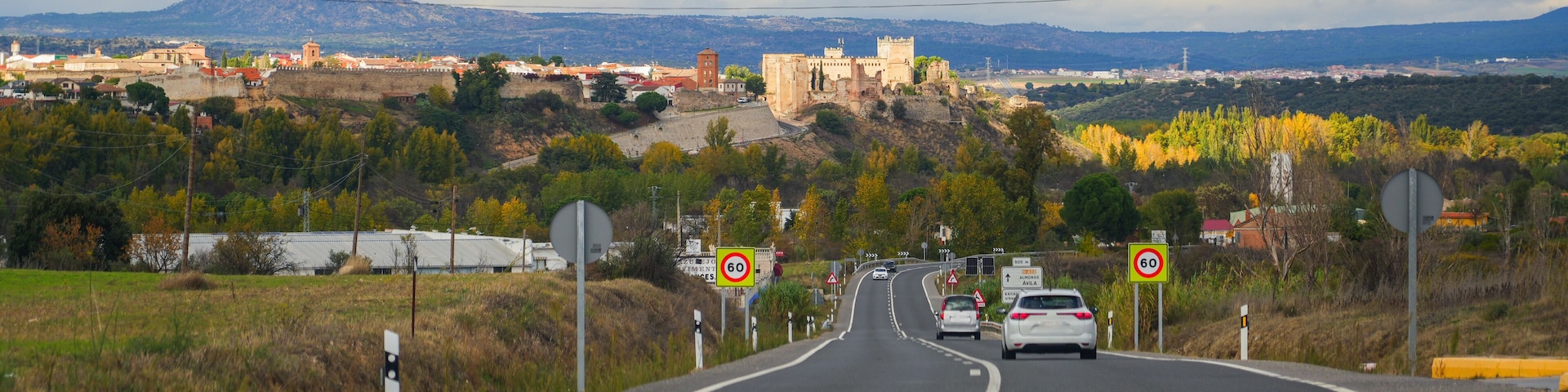 Carretera que conduce a un castillo histórico en una colina, con un letrero de límite de velocidad de 60 km/h y un indicador de un pueblo cercano.