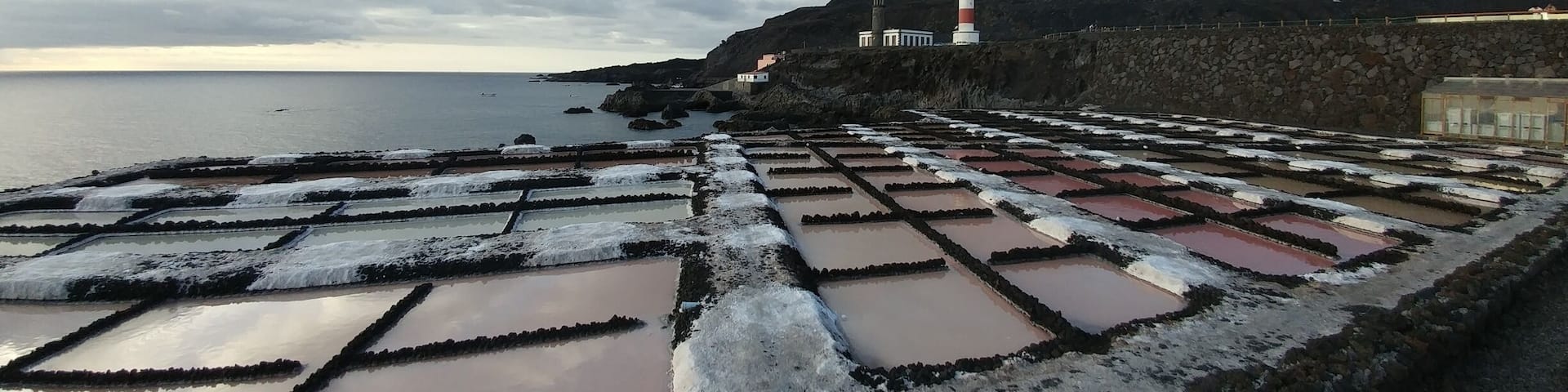 A coastal landscape with a lighthouse, terraced salt pans, and a stone wall.