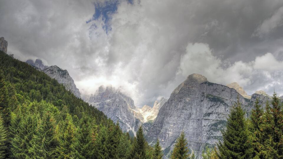 A mountainous landscape with dense forests and rugged peaks under a cloudy sky.