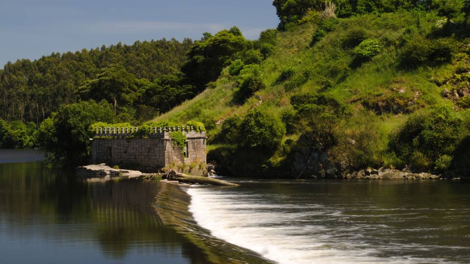 A river flowing past a stone structure with a fence, surrounded by lush greenery and hills.