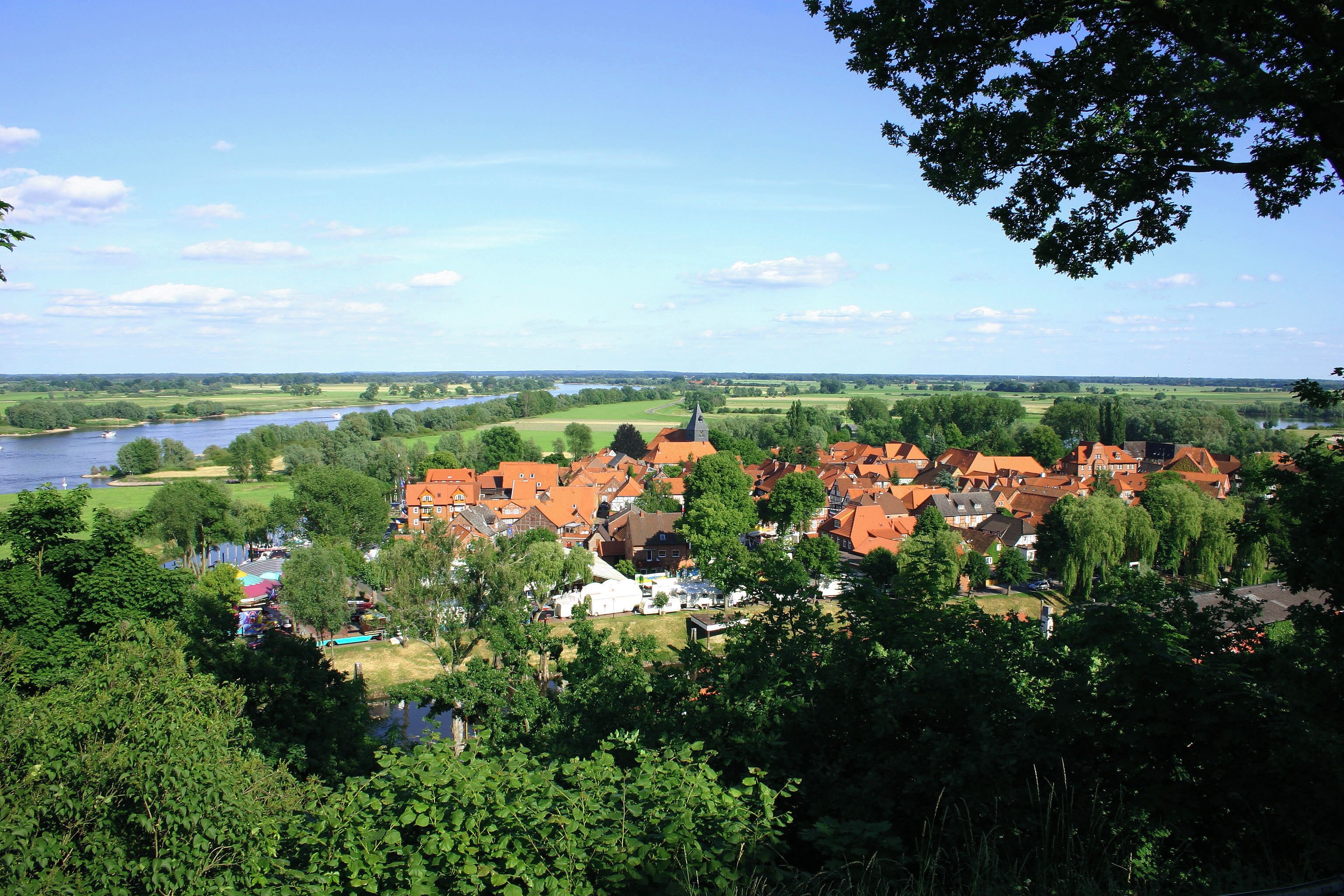 A riverside town with red-roofed houses, a church steeple, and lush greenery.