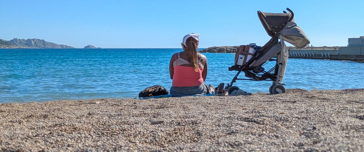 A person sitting on a pebbled beach with a stroller and a bag, near a calm sea and rocky coastline.