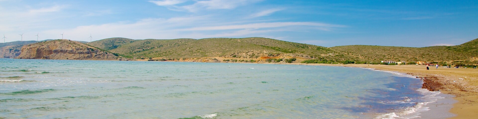 Una playa con agua cristalina, orilla arenosa y aerogeneradores al fondo.