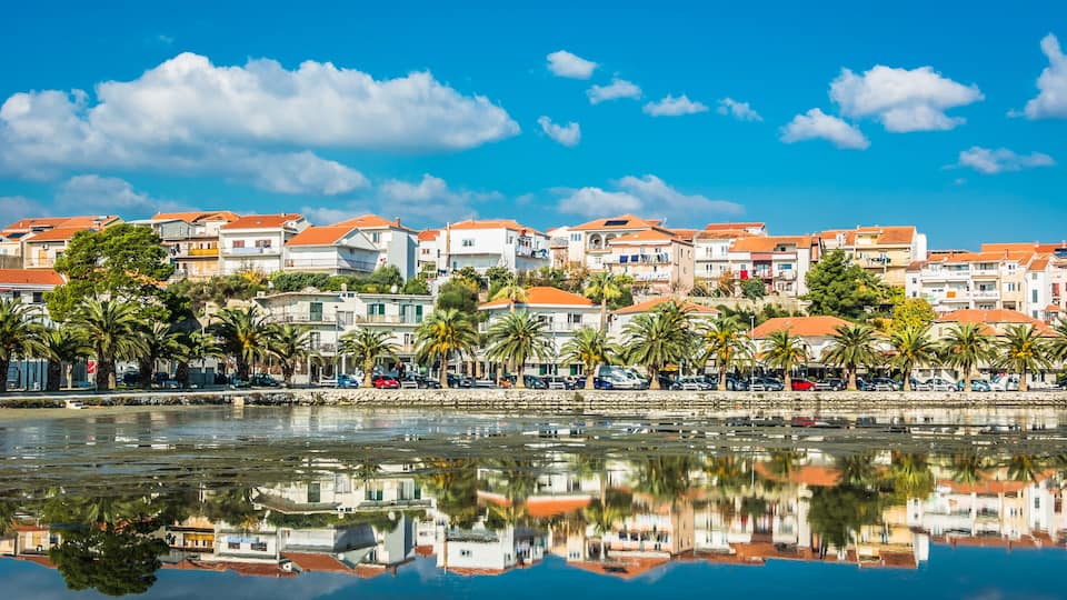 A coastal town with buildings, palm trees, and a calm sea.