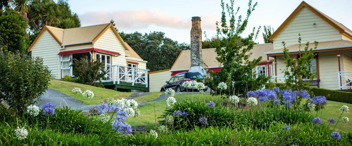 A row of two-story houses with red and white trim, surrounded by lush greenery and flowering plants.