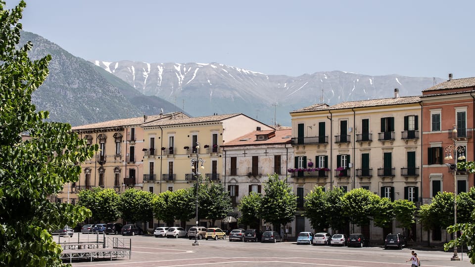 A town square with parked cars, trees, and buildings with balconies. Snow-capped mountains are in the background.