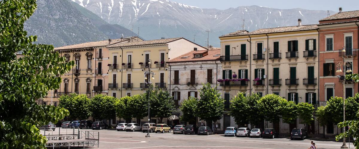 A town square with parked cars, trees, and buildings with balconies. Snow-capped mountains are in the background.