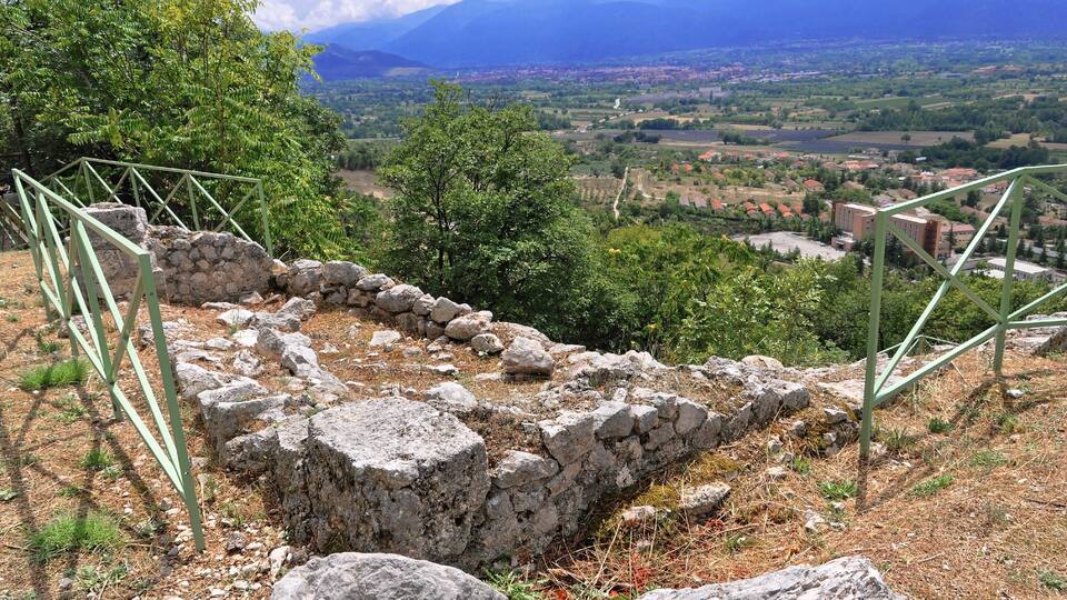 Ancient ruins with stone walls and metal railings, surrounded by trees and a mountainous landscape in the background.