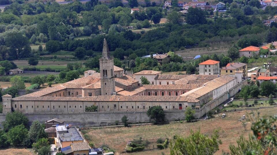 A fortified complex with a central tower, surrounded by buildings with red-tiled roofs, set in a rural landscape with greenery and distant houses.