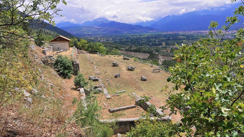 A hillside with ancient ruins, a small building, and a distant mountain range.