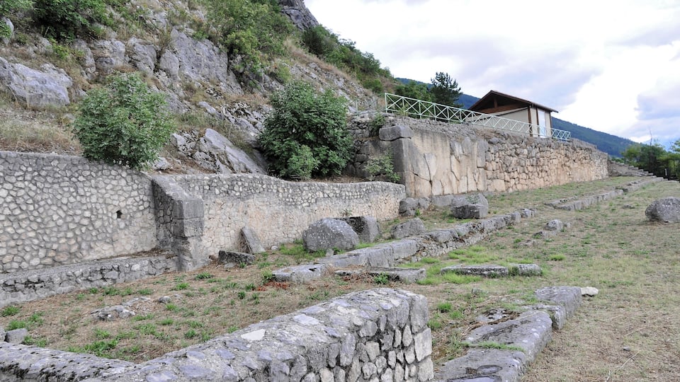 Ancient stone ruins with a stone wall and a small building on a hillside.