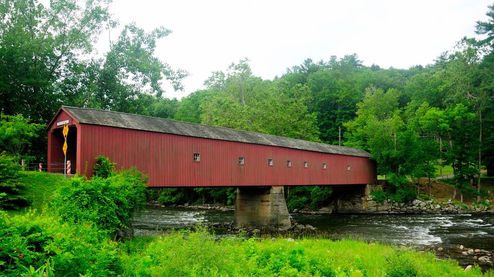 A red covered bridge over a river, surrounded by lush greenery and trees.