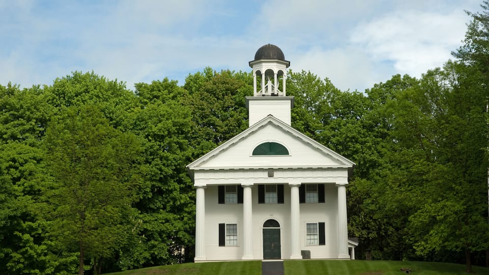 A white building with a central dome and columns, surrounded by lush green trees.