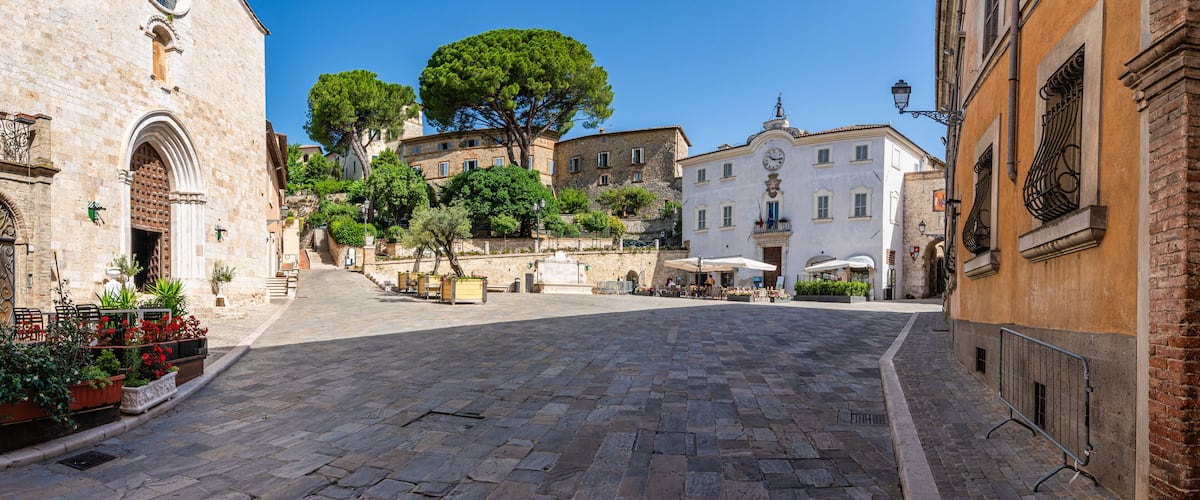 A cobblestone street with historic buildings, a large tree, and outdoor seating areas.