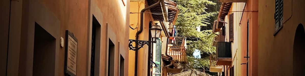 A narrow street with buildings on either side, a sign for 'SP Stazione Di Posta San Gemini', and a car parked at the end.