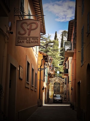 A narrow street with buildings on either side, a sign for 'SP Stazione Di Posta San Gemini', and a car parked at the end.