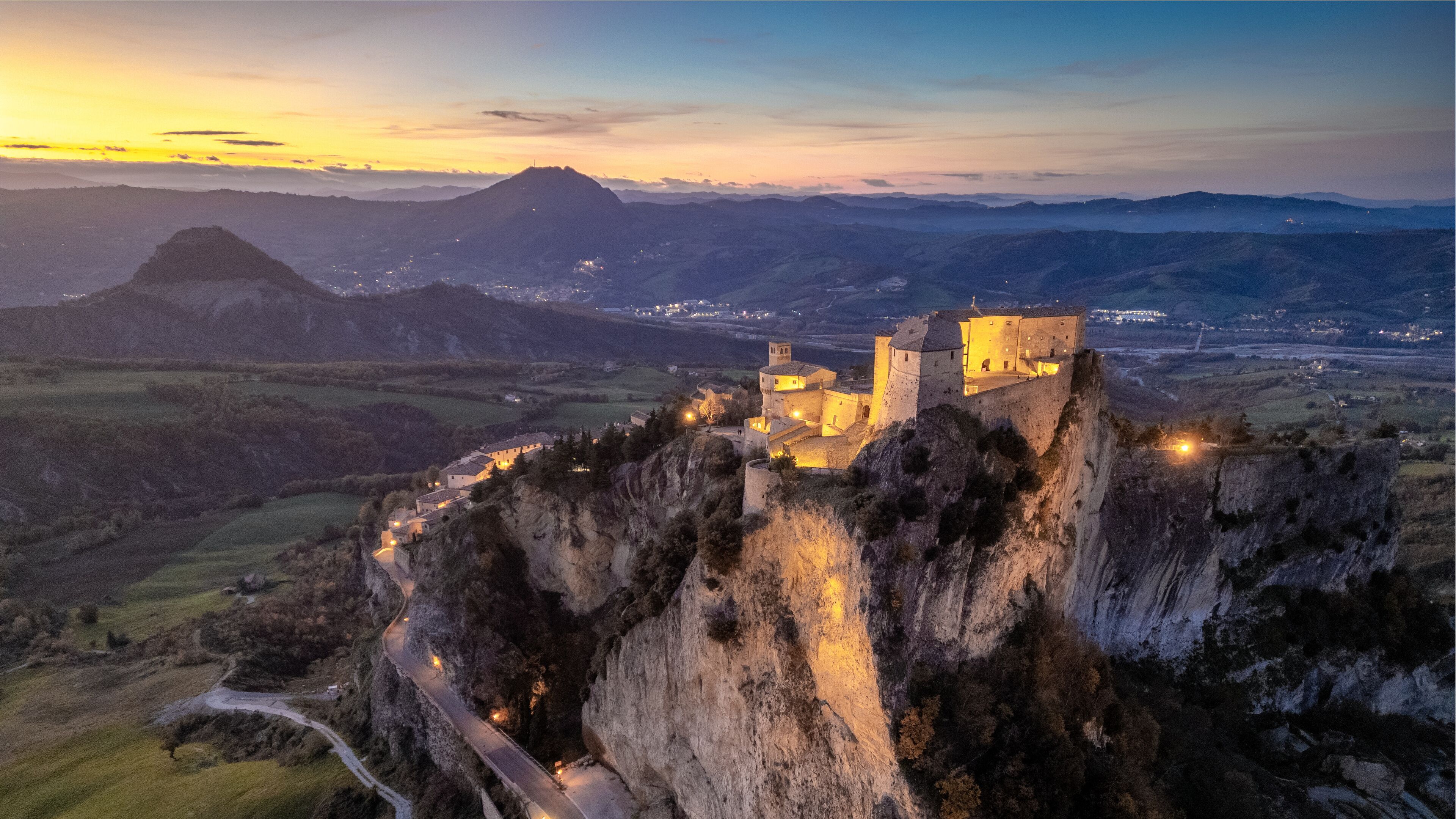 Un castillo medieval se alza sobre un acantilado con vistas a un valle al anochecer.