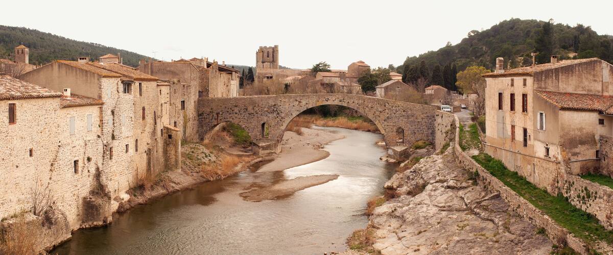 A stone bridge over a river in a historic town with old buildings and a hill in the background.