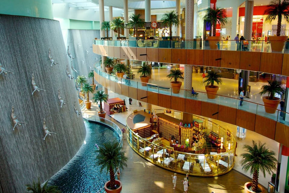 A multi-level shopping mall with a central atrium, palm trees, and a water feature.
