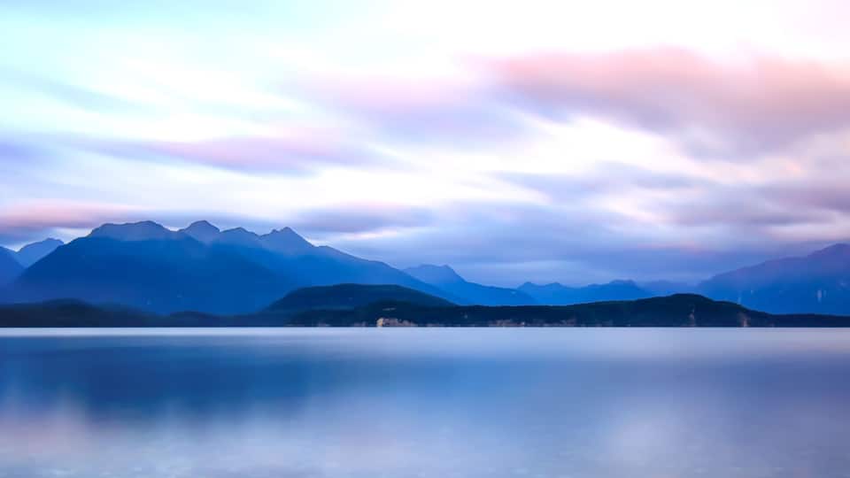A mountain range reflected in a calm lake with a sky featuring soft pink and blue hues.