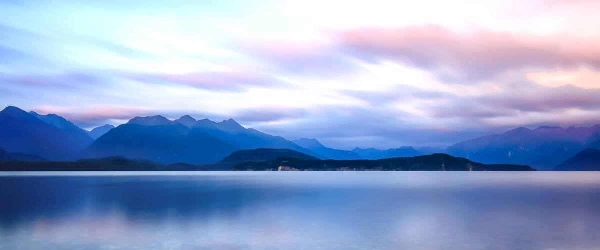 A mountain range reflected in a calm lake with a sky featuring soft pink and blue hues.