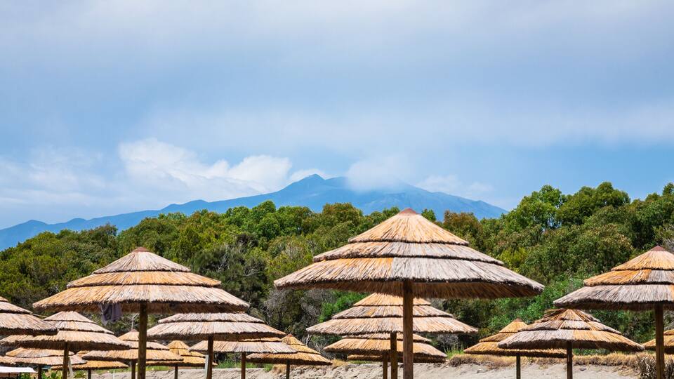 A beach with thatched umbrellas and a mountain in the background.