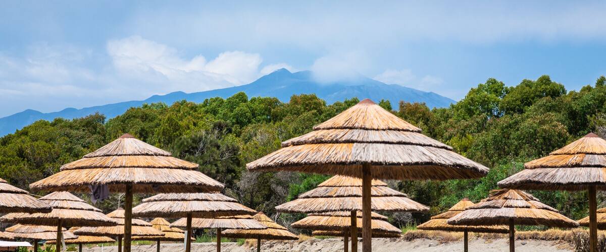 A beach with thatched umbrellas and a mountain in the background.