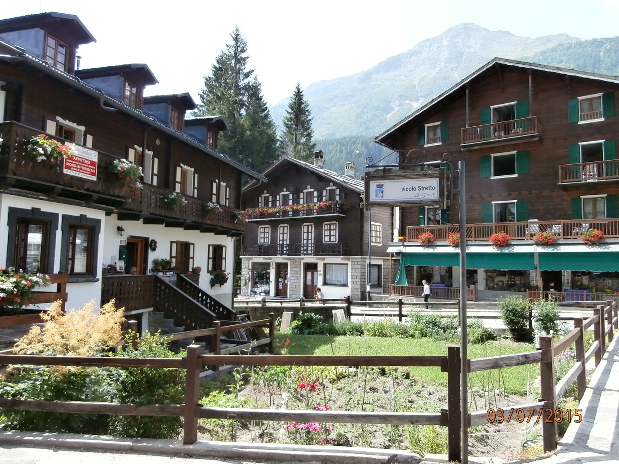 A traditional wooden building with balconies, a sign reading "vicolo Stretta," and a mountainous backdrop.