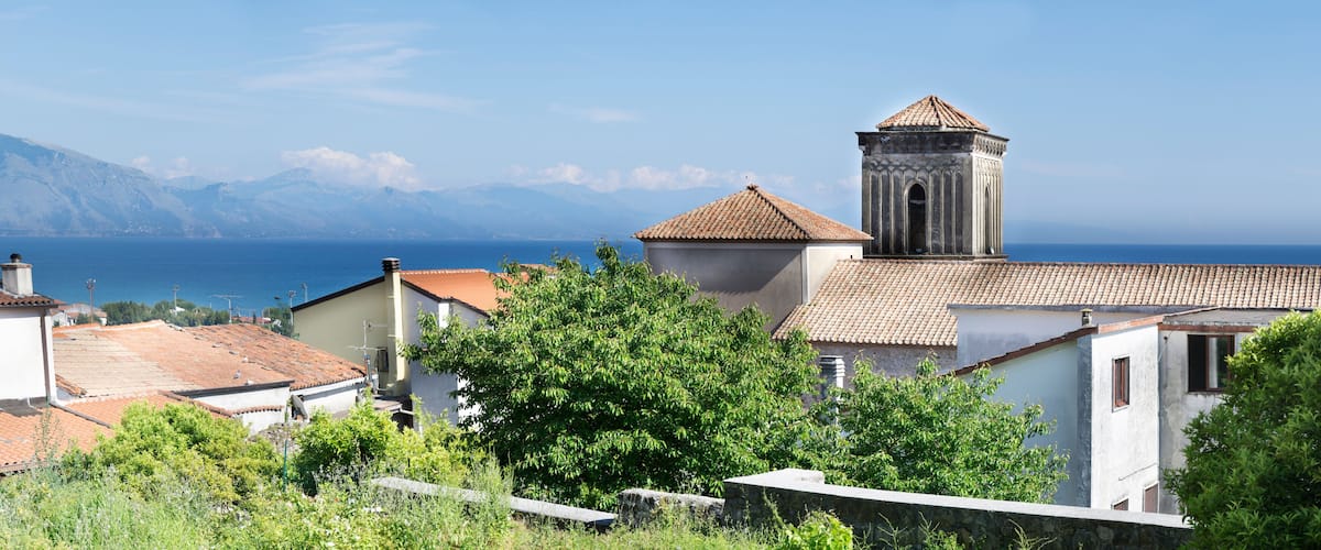 A coastal village with terracotta-roofed buildings, a bell tower, and a backdrop of mountains and the sea.