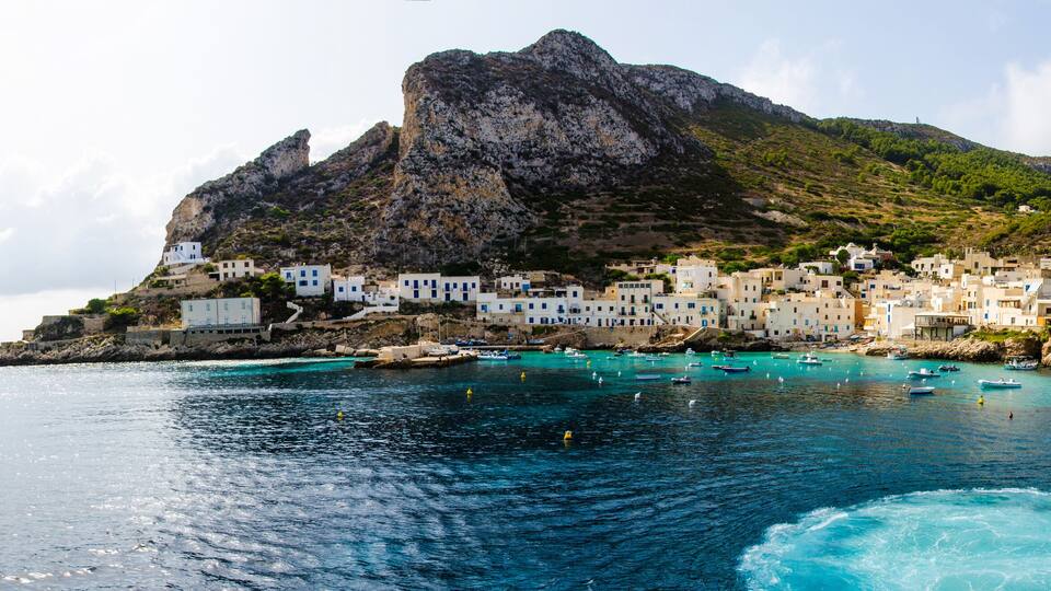 A coastal town with white buildings, a rocky mountain, and turquoise waters.