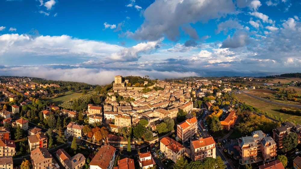 Une vue panoramique dâune ville historique, avec un chĂąteau imposant qui domine une colline, entourĂ©e de bĂątiments rĂ©sidentiels et dâun mĂ©lange de champs verdoyants et de routes.