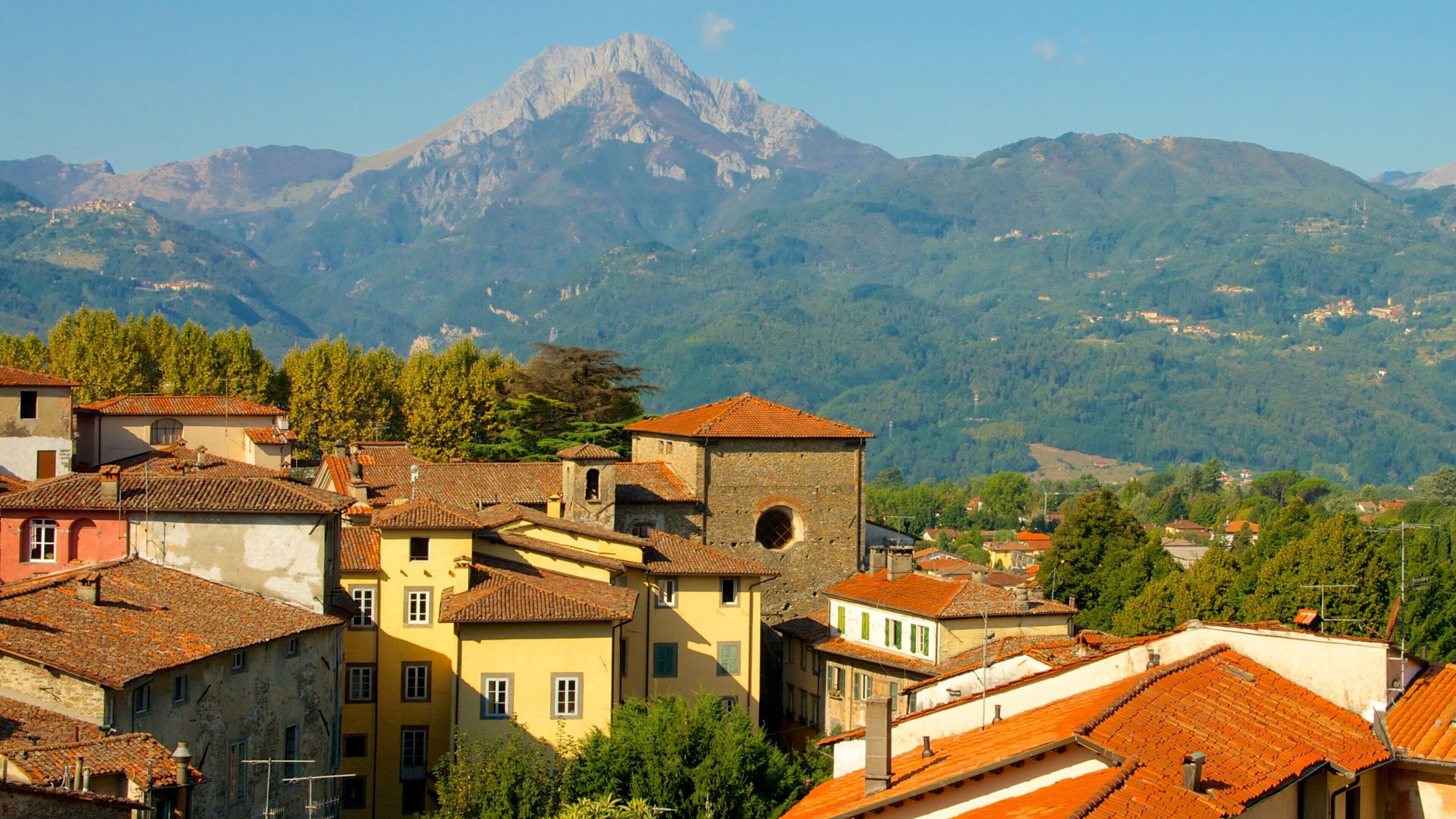 A town with terracotta-roofed buildings, a prominent tower, and a mountain range in the background.