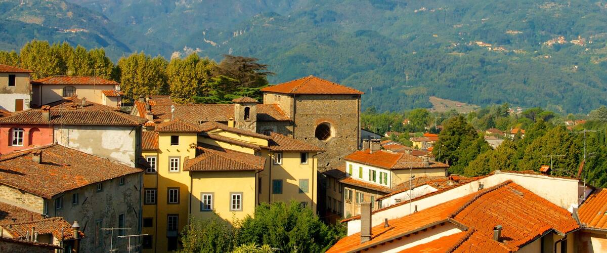 A town with terracotta-roofed buildings, a prominent tower, and a mountain range in the background.