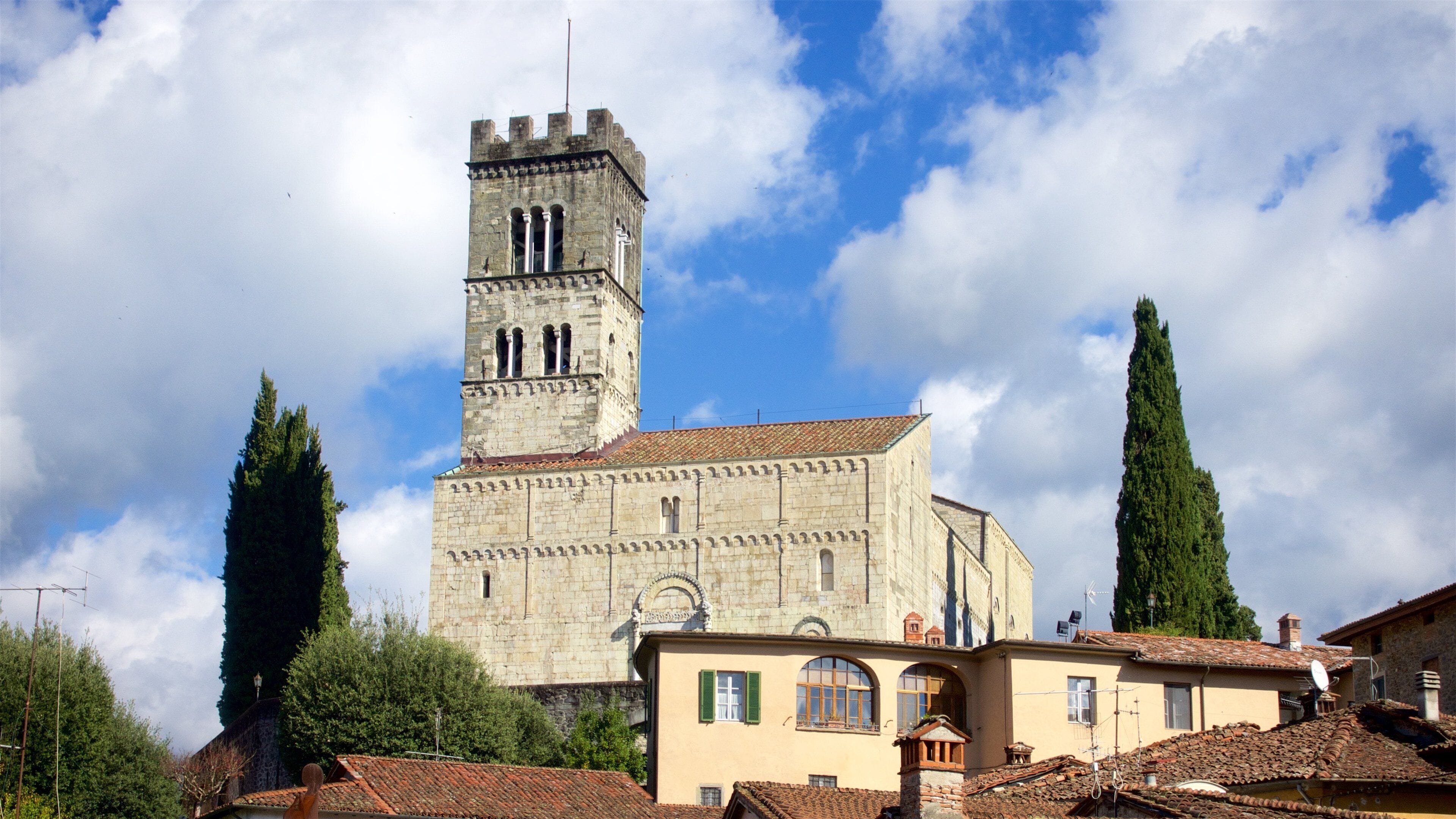 A historic stone church with a tall bell tower, surrounded by traditional buildings and tall cypress trees.
