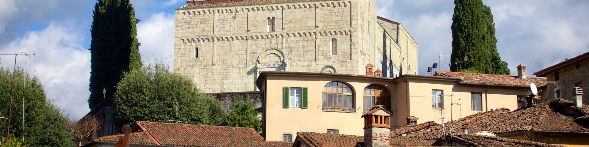 A historic stone church with a tall bell tower, surrounded by traditional buildings and tall cypress trees.
